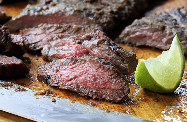 Sliced flank steak on a metal sheet pan with lime wedges.